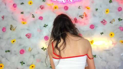 Media: Video of a woman with long black hair, wearing a white tank top with red trim, facing away from a floral wallpaper backdrop adorned with butterflies, sunflowers, and pink roses.