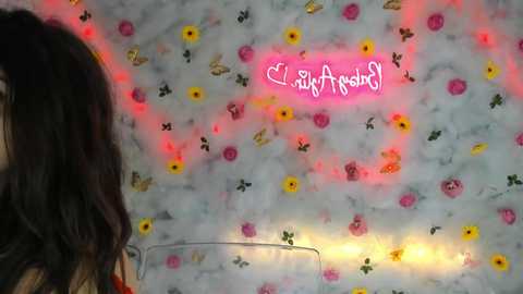 Media: A video of a woman with long, dark hair, standing against a floral wallpaper with a pink neon heart sign. The background features yellow, red, and white flowers.