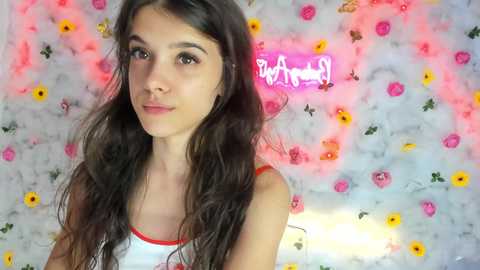 Video of a young, light-skinned Latina woman with long, wavy brown hair and dark eyes, wearing a white tank top with red trim, standing in front of a floral backdrop with pink and yellow flowers.