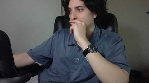 Media: A video of a young man with curly dark hair, wearing a blue button-up shirt, sitting in a black office chair, with a serious expression, hand to his chin, indoors.