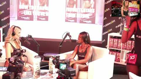 Media: Video of two women in lingerie, one with long brown hair, the other with short blonde hair, seated on a white couch. Background features promotional posters and studio equipment.