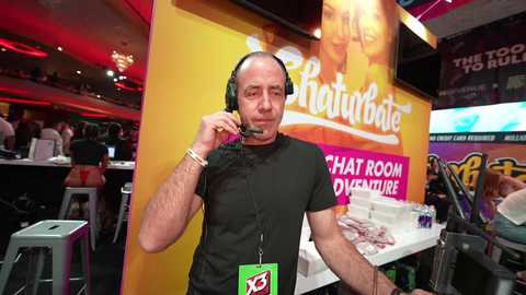 Media: Video of a middle-aged bald man in a black t-shirt, wearing headphones and a lanyard, standing in a lively, colorful bar with red and yellow walls, posters, and a crowded background.