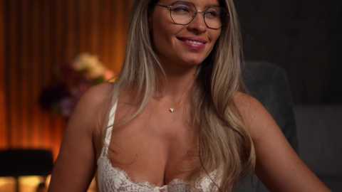 Media: Video of a smiling, fair-skinned woman with long, straight blonde hair, wearing glasses, a lacy white bra, and a delicate gold necklace. Background shows a blurred, dimly lit room with a warm, amber glow.