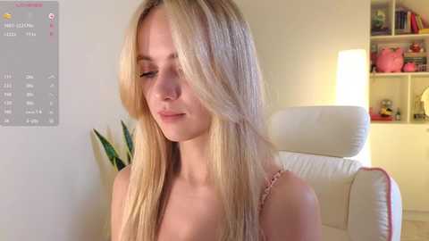 Media: Video of a young, fair-skinned, blonde woman with straight hair, wearing a pink camisole, sitting in a white armchair in a brightly lit, modern living room with a green plant and bookshelves in the background.