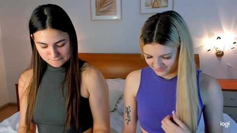 Media: Video of two young women with long, straight hair; one brunette in a black crop top, one blonde in a purple crop top, standing in a modern bedroom.