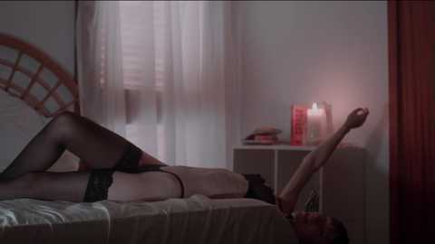 Media: A dimly lit video depicts a woman in black lingerie and stockings lying on a bed, stretching her arms towards a lit candle on a white nightstand in a cozy, softly lit bedroom.