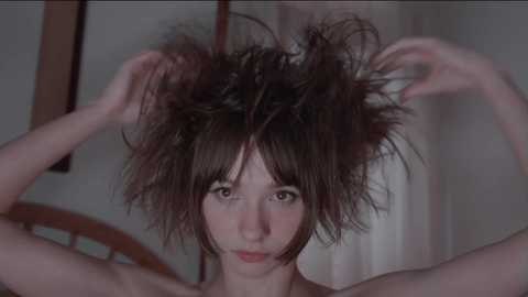 Media: Video of a young, pale-skinned woman with short, messy, dark brown hair, holding her hair up with both hands. Background features a wooden chair and white curtain.