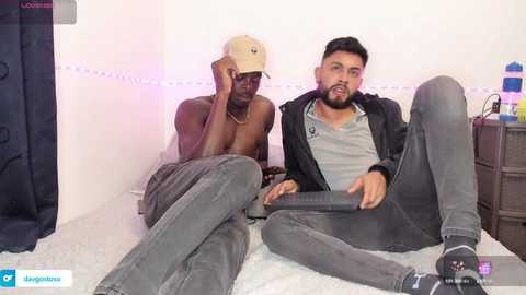 Media: Video of two men: a shirtless Black man with a cap and grey pants, and a bearded Middle Eastern man in a grey hoodie and jeans, sitting on a bed with white sheets, in a dimly lit room.