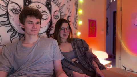 Video of two young people with pale skin and light hair in a cozy room with sun and moon tapestry, neon lights, and plaid blanket.