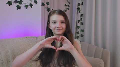 Media: Video of a young Caucasian woman with long, dark hair forming a heart shape with her hands. She has fair skin and is smiling. Background includes a beige couch, green ivy garland, and purple lighting.