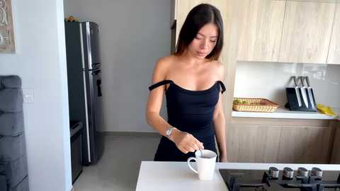 Video of an Asian woman with long black hair, wearing a black off-shoulder dress, pouring coffee in a kitchen with modern wooden cabinets and a black refrigerator.