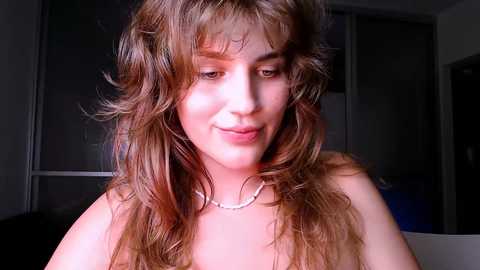 Media: Video of a young woman with wavy, shoulder-length brown hair, wearing a pearl necklace, smiling softly, indoors with dark background.