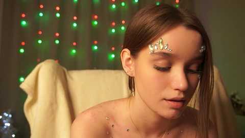 Video of a young, fair-skinned woman with straight, light brown hair, wearing silver leaf-shaped headpieces and delicate earrings, sitting on a beige cushioned chair in a softly lit room with festive green and red fairy lights in the background.