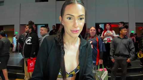 Media: Video of a young woman with dark hair in braids, wearing a low-cut yellow bikini top and black jacket, posing at an indoor event with people in casual attire in the background.