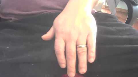 Media: Video of a light-skinned hand wearing a silver ring resting on a black car seat cushion. The background features a blurred interior scene with a purple shirt and a wooden floor visible.