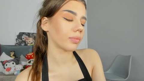 Media: Video of a young woman with light skin and brown hair, wearing black tank top, applying makeup in a minimalist room with grey walls, gray chair, and decorative pillows.