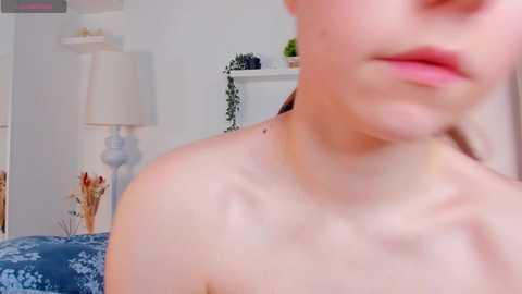 Media: Video of a fair-skinned woman with light pink lips, wearing no top, showing a sunburn on her shoulders. Background includes a white shelf with plants, a blue-patterned blanket, and a lamp.