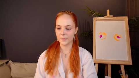 Video of a pale-skinned, red-haired woman in a white robe, sitting in front of an easel with a floral painting. Background features a dark wall and greenery.