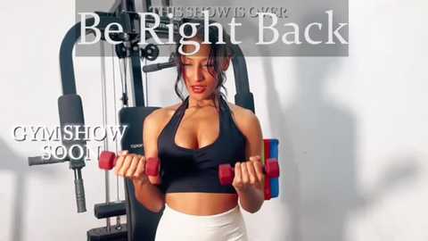 Media: Video of a muscular, fit woman with long black hair in a black sports bra and white leggings, lifting red dumbbells in a gym, with text overlay \"Be Right Back\" and \"GYMSHOW 500.\