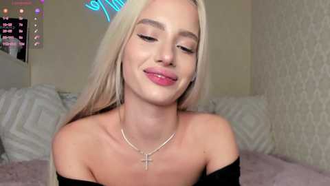 Video of a young, smiling woman with long, straight platinum blonde hair, fair skin, wearing a black off-shoulder top, and a silver cross necklace. Background shows a cozy, white-patterned room with a bed and neon lights.