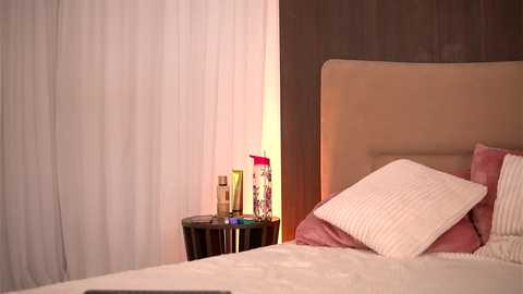 Media: Video of a minimalist bedroom with a beige upholstered headboard, white curtains, a bedside table holding a red candle and decorative items, and a neatly made bed with white and pink pillows.