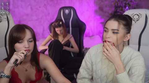 Video of two women, one with brown hair and a red top, the other with brunette hair and a beige sweater, eating in a dimly lit room with purple lighting, behind a black gaming chair.