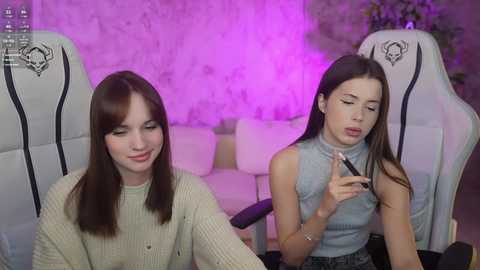 Media: Video of two young women in gaming chairs, one applying makeup, the other smiling, in a softly lit room with purple accents.