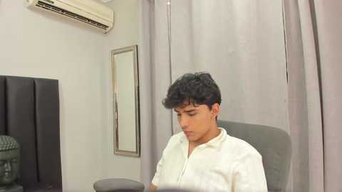 Media: Video of a young man with short dark hair, wearing a white shirt, sitting on a grey chair in a sterile room with grey curtains, air conditioner, and a black statue in the background.