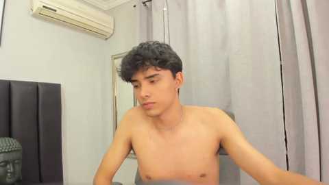 Media: Video of a shirtless, slim, young Latino man with curly black hair, sitting on a bed, wearing only gray boxer shorts. Background shows light walls, air conditioner, and gray curtains.