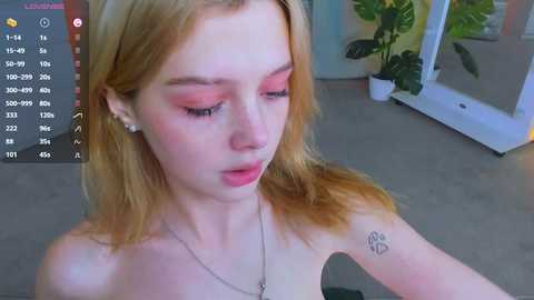 Media: Video of a young woman with fair skin, blonde hair, and pink makeup, wearing a silver necklace and a small tattoo on her right arm. She is topless, indoors with a mirror and green plant in the background.