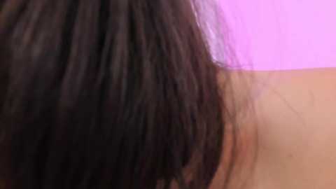 Media: A close-up video of a person's shoulder with long, dark brown hair partially covering the face. The background is a soft pink hue, providing a contrasting and vibrant backdrop. The image is grainy and lacks detailed facial features.