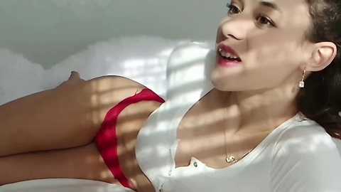 Media: Video of an East Asian woman with fair skin, lying on a white bed. She wears a white crop top and red panties, casting shadow patterns on her body. Sunlight filters through blinds, creating a soft, intimate ambiance.