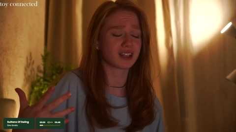 Media: Video of a fair-skinned redhead woman with long hair, wearing a light blue shirt, mouth open in distress, indoors with warm lighting, blurred background.