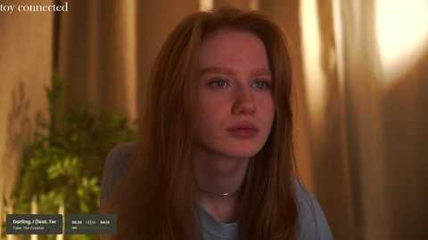 Media: A video of a fair-skinned, red-haired woman with blue eyes, wearing a light blue top, indoors with warm lighting and a blurred potted plant in the background.