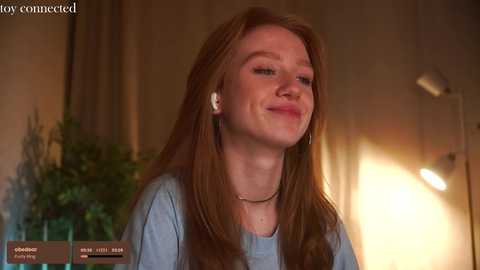 Media: Video of a fair-skinned, red-haired woman with long hair, wearing a light blue shirt, smiling slightly, indoors with a potted plant and lamp in the background.