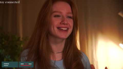 Media: Video of a smiling young woman with long red hair, wearing a light blue top, in a warmly lit room with a fireplace.