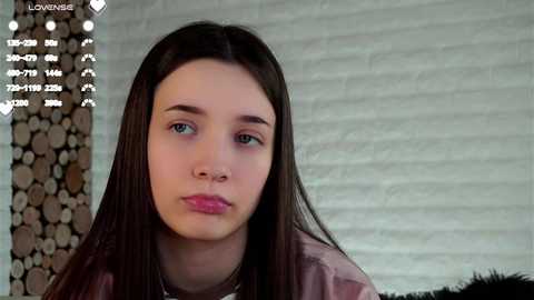 Media: Video of a young woman with long dark hair, fair skin, and blue eyes, wearing a pink blouse, set against a rustic brick wall with a woodpile to the left.