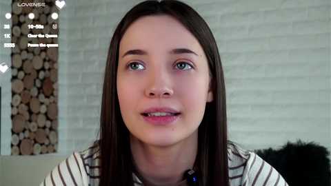 Video of a young woman with straight, dark brown hair and light skin, wearing a striped shirt. She has a neutral expression and is indoors with a brick wall background and a pile of wood logs.