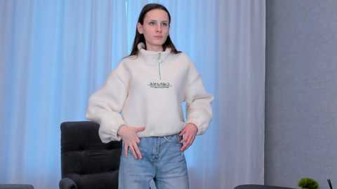 Media: Video of a young woman with fair skin and straight brown hair, wearing a white \"Champion\" hoodie and light blue jeans, standing confidently indoors with a black chair and light blue curtains in the background.