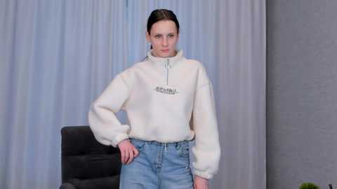 Media: Video of a young, fair-skinned woman with straight, dark hair, wearing a cream-colored, fuzzy sweater with a small black logo, paired with high-waisted, light blue jeans. She stands confidently in front of light gray and white curtains, with a black chair partially visible behind her.