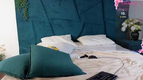 Media: Video of a modern bedroom with teal velvet headboard, white pillows, green throw pillows, and black keyboard on a beige bedspread.
