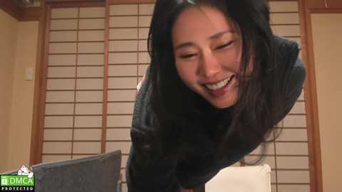 Media: A video of a smiling Asian woman with long black hair, wearing a black jacket, leaning over a table, in a room with beige walls and wooden shoji screens.