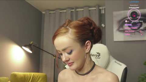 Media: Video of a fair-skinned woman with red hair styled in a bun, wearing a black choker necklace and makeup, seated in a grey office chair with a yellow cushion, against a grey wall, with a lit desk lamp and a framed eye image in the background.