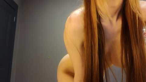 Media: A video of a nude woman with long, straight, auburn hair, leaning forward, her upper body visible, with a gray wall and door in the background.