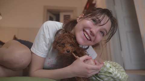 Media: Video of a young woman with wet hair, wearing a white t-shirt, smiling and holding a wet, curly-haired dog. Background shows a blurred indoor setting with beige walls and a green bedspread.