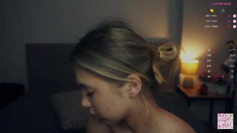 Media: Video of a woman with light skin, blonde hair in a bun, wearing a sheer top, sitting on a bed. The room is dimly lit with a warm lamp on a nightstand.