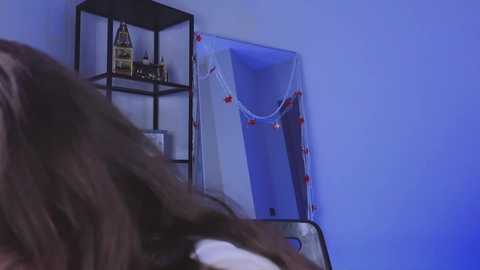 Media: A video of a young girl with long, brown hair in a blue room, taking a selfie in front of a white, empty room with a string of red lights. A small black shelf with toys and books is visible in the background.