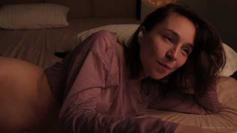 Media: Video of a woman with shoulder-length brown hair, wearing a loose purple shirt, lying on a bed, partially covered by white and beige sheets, in a dimly lit bedroom.