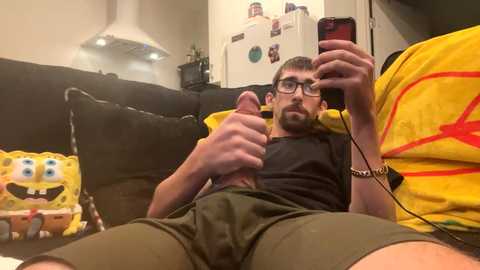 Media: Video of a bearded man with glasses, giving a thumbs-up while holding a phone, wearing a black shirt and green shorts. Background includes a black couch with SpongeBob SquarePants and a yellow Pikachu plush toy.