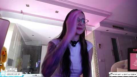 Media: Video of a young woman with long brown hair, wearing glasses, a white blouse, and a black bow tie, standing in a modern room with purple lighting and a mirror.